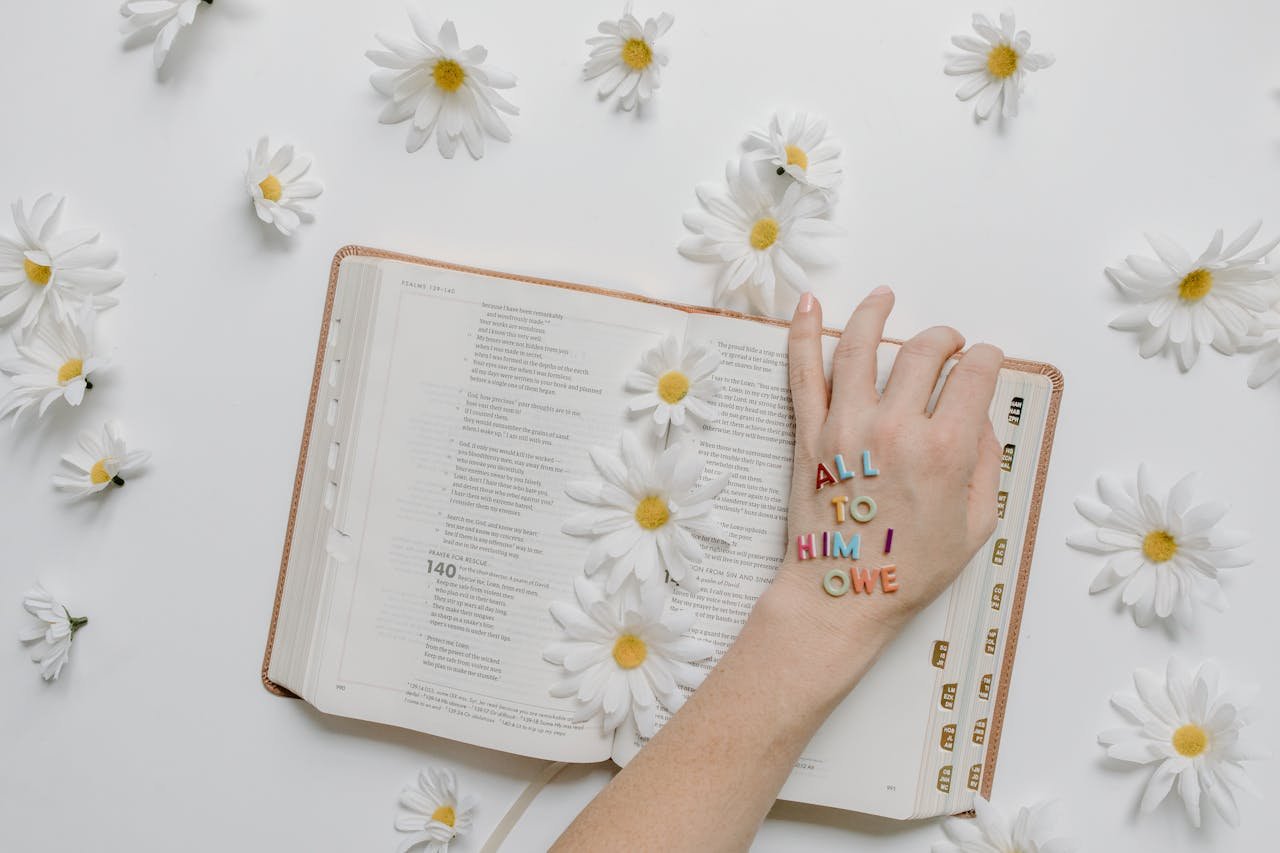 Open Bible adorned with daisies and letters spelling 'ALL TO HIM I OWE', emphasizing faith and inspiration.